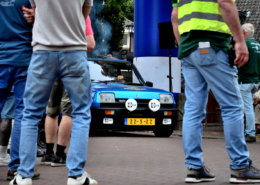 front left angle view of a sky blue classic Renault Turbo car in the background, spectators and crew standing on a red brick road in the foreground. vantage point through the legs of the spectators